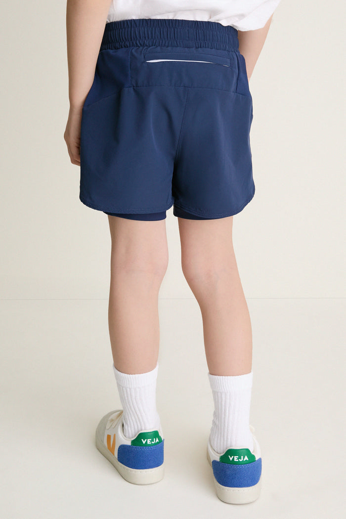 Back view of a child wearing classic navy-blue Before Sunrise Blue Cardio Short with elastic waistband, visible liners, and a zippered pocket, paired with stylish white sneakers.