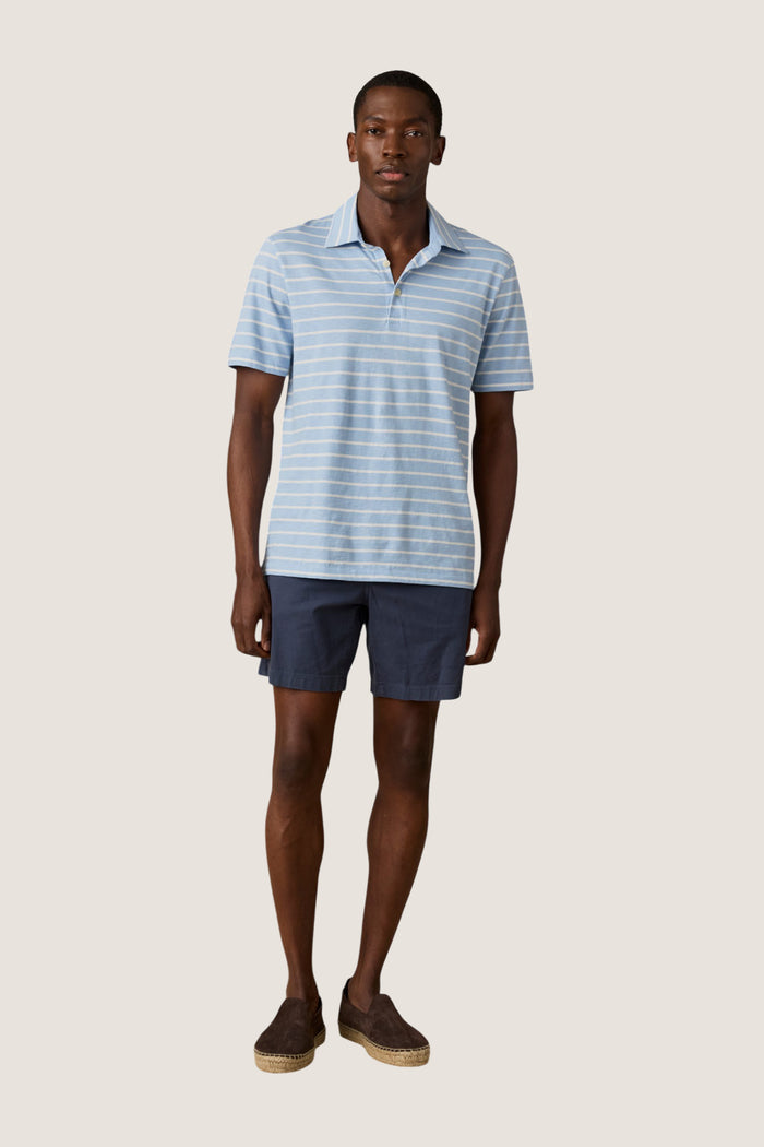 Model wearing the Bermuda Sail Stripe Sunwashed Tee Shirt Polo, a classic, effortless light-blue polo with white stripes, paired with navy shorts and dark-brown espadrilles.