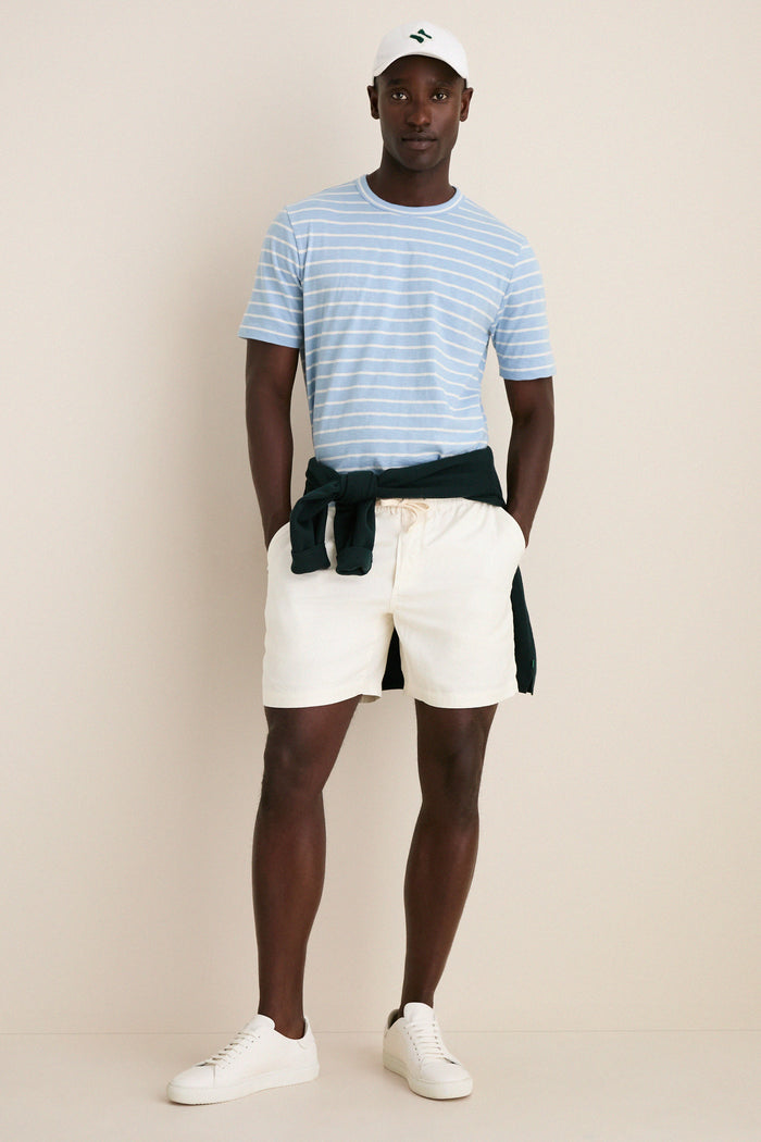 Person wearing a Bermuda Sail Stripe Sunwashed Tee, exuding effortless, classic style with nautical stripes. Paired with off-white shorts and a dark green sweatshirt, embodying high-end, fun fashion.