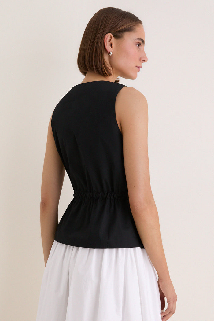 Woman in a sleeveless, classic Black Cotton Poplin Francoise Top with high neckline and cinched waist, exuding effortless sophistication against a simple backdrop.