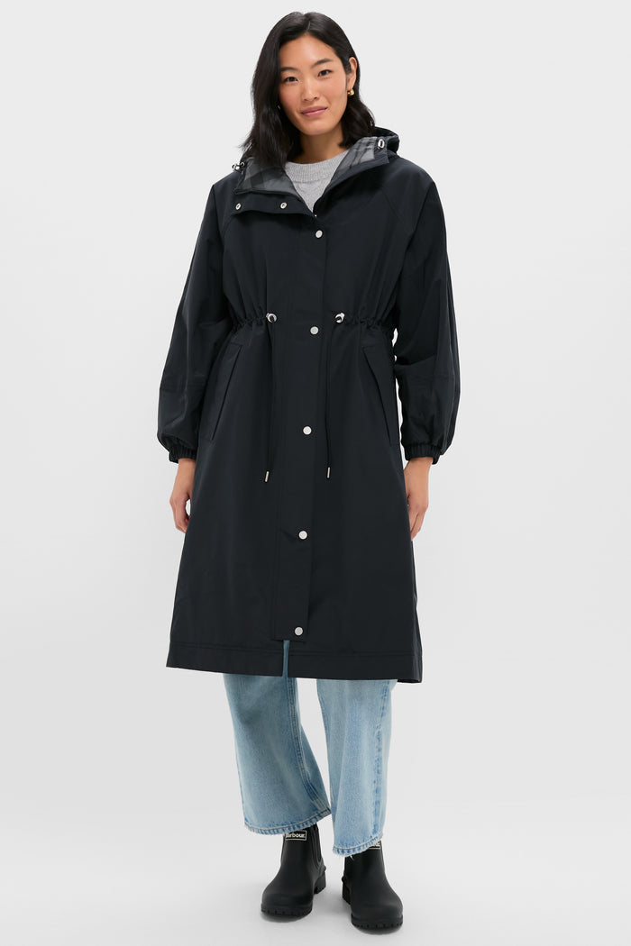A woman in a Barbour long black waterproof parka with drawstring waist, light blue jeans, and black boots stands against a plain light background.