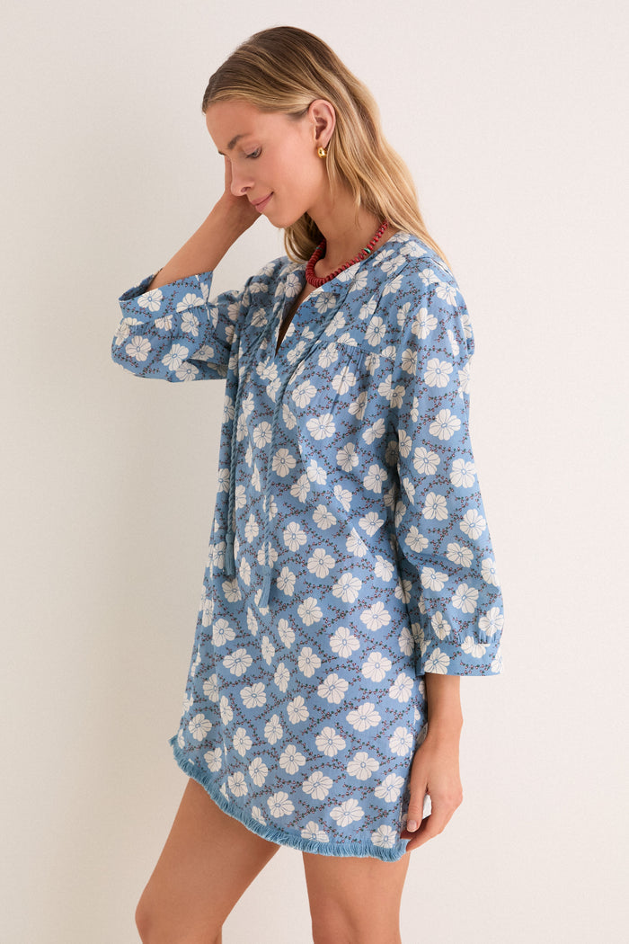 Model in Bloom Lagoon Bondi Coverup, a classic, effortless tunic with floral pattern, tassel neckties, and fringed hem. Accessorized with red necklace, gold hoops.