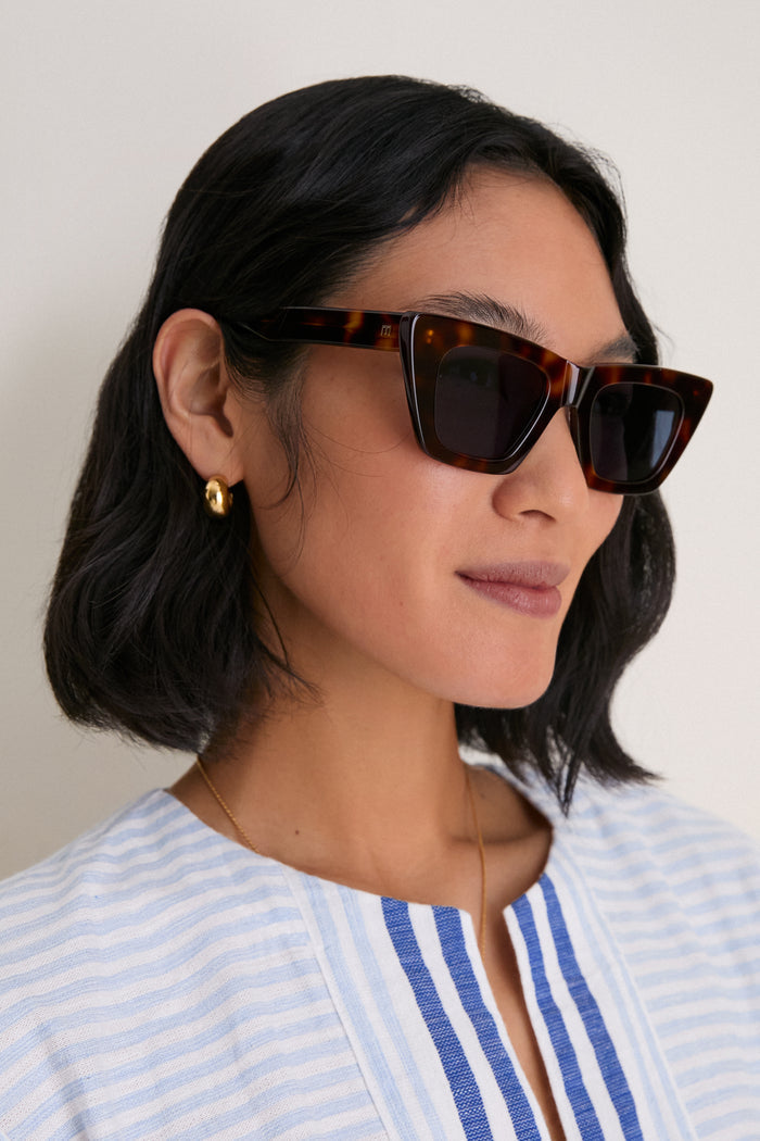 Woman in tortoiseshell cat-eye sunglasses with blue lenses, exuding classic, effortless style. Accompanied by gold hoop earrings and striped blouse for a high-end, fun look.