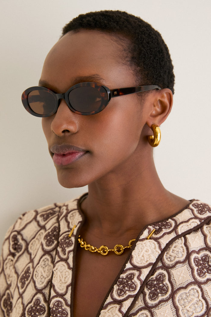 Person wearing Brown Tortoise Lyric Leigh Sunglasses, showcasing their classic oval shape. Effortlessly paired with chunky gold accessories, embodying a fun, high-end style.