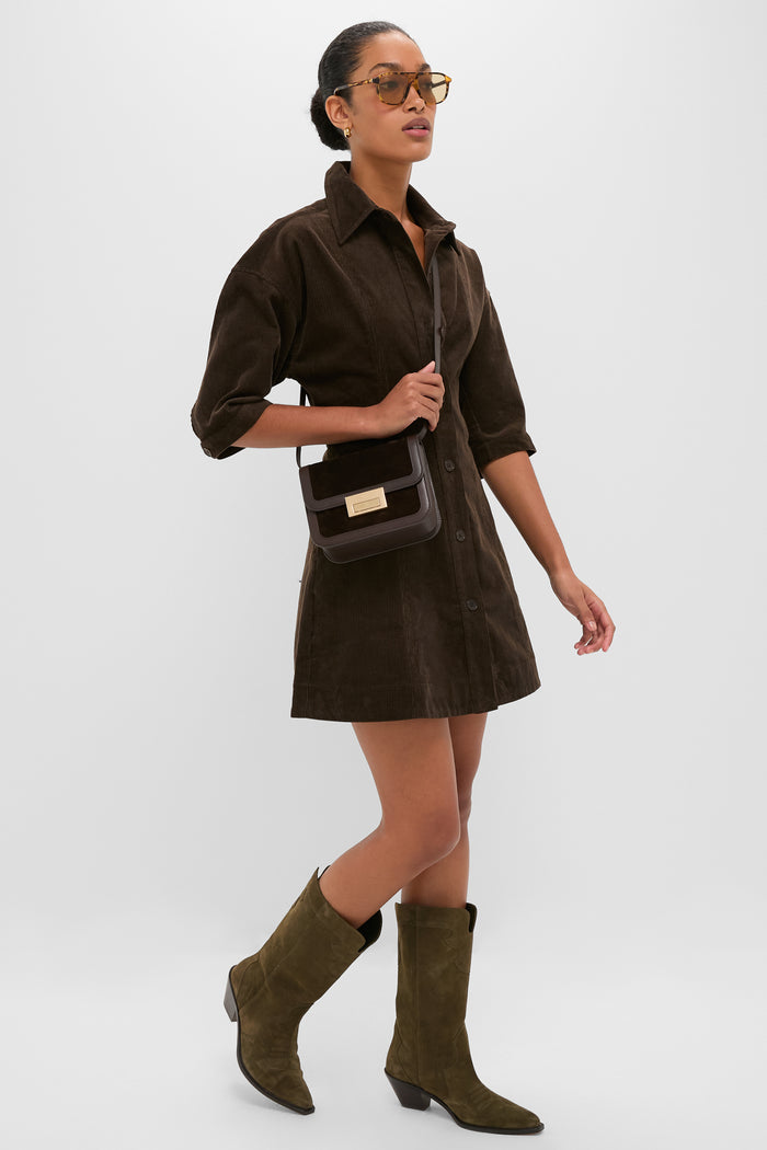 Woman wearing the Carob Luz Mini Dress, a classic corduroy shirt-dress with lantern sleeves, paired with tortoiseshell sunglasses, dark leather bag, and olive suede boots; effortlessly high-end.