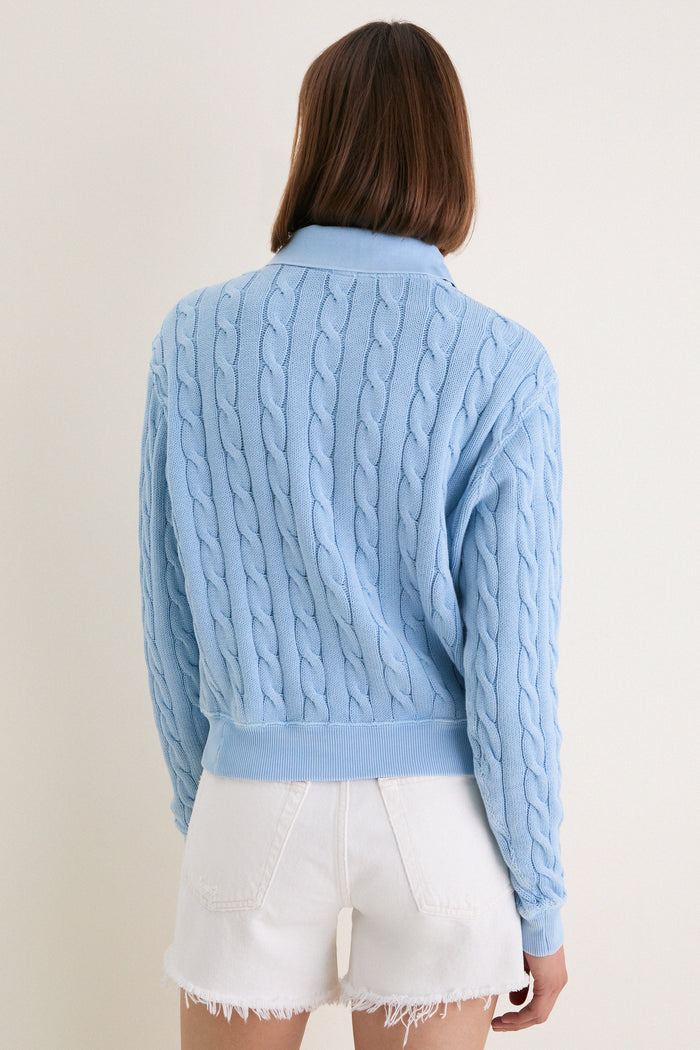 Person wearing Chambray Blue Cotton Knit Polo with classic cable-knit design, polo collar, and ribbed details, paired with high-waisted white denim shorts for an effortless, high-end look.