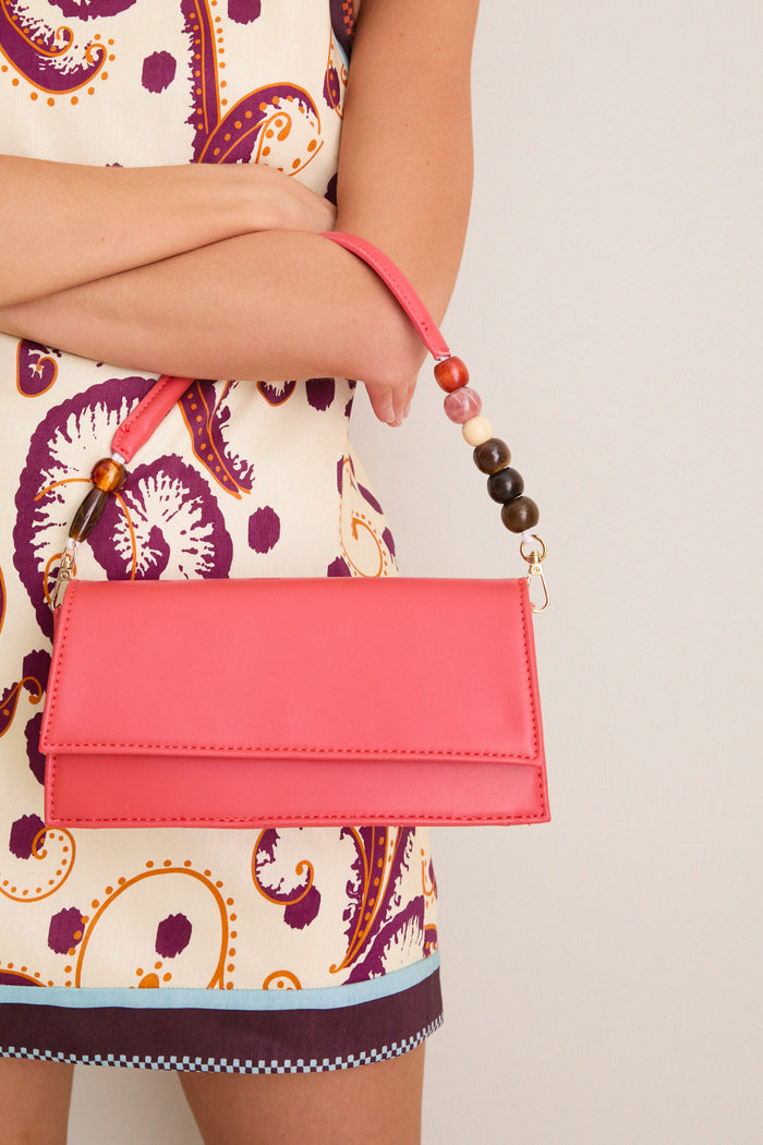 Person holding the Coral Brooks Crossbody, a classic coral-pink envelope handbag with a beaded handle and gold-tone hardware, exuding effortless, high-end style and fun versatility.
