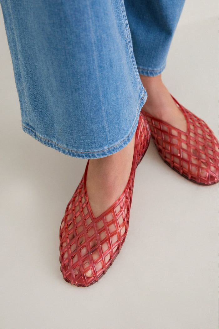 Lower legs wearing Coral Jam Jellys, red lattice-style flats made of semi-transparent plastic, paired with light blue cropped jeans, showcasing classic, effortless, and fun high-end style.
