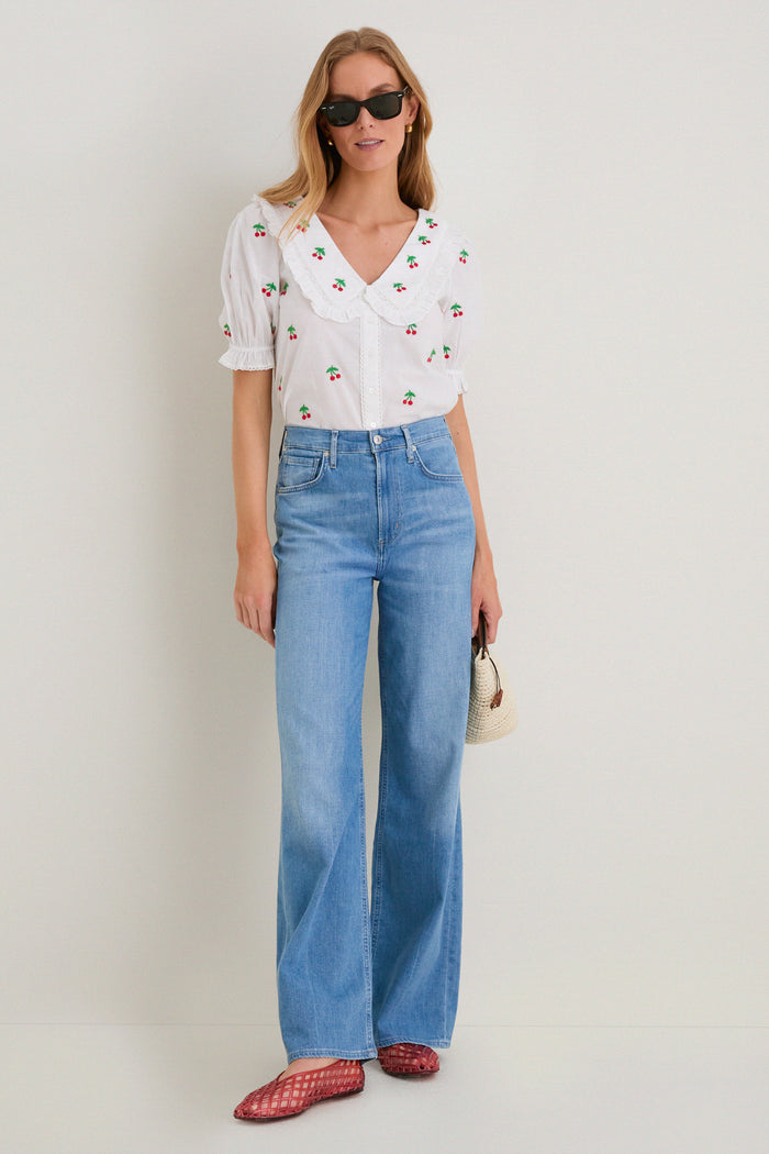 Woman in Coral Jam Jellys, a classic, effortless, and fun look with a cherry-print blouse, light-blue flared jeans, red flats, and a woven straw bag.