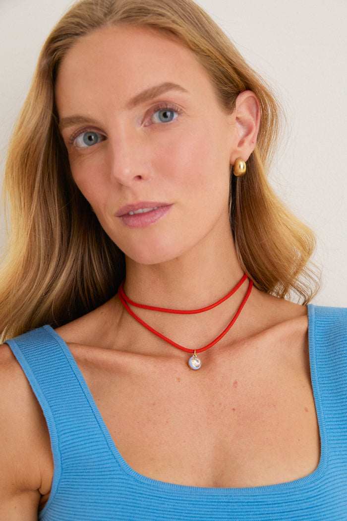 A classic, effortless Coral Red Necklace Cord with Blue Bezel Charm adds a fun, high-end touch, worn by a person with light hair, gold earrings, and a blue tank top.