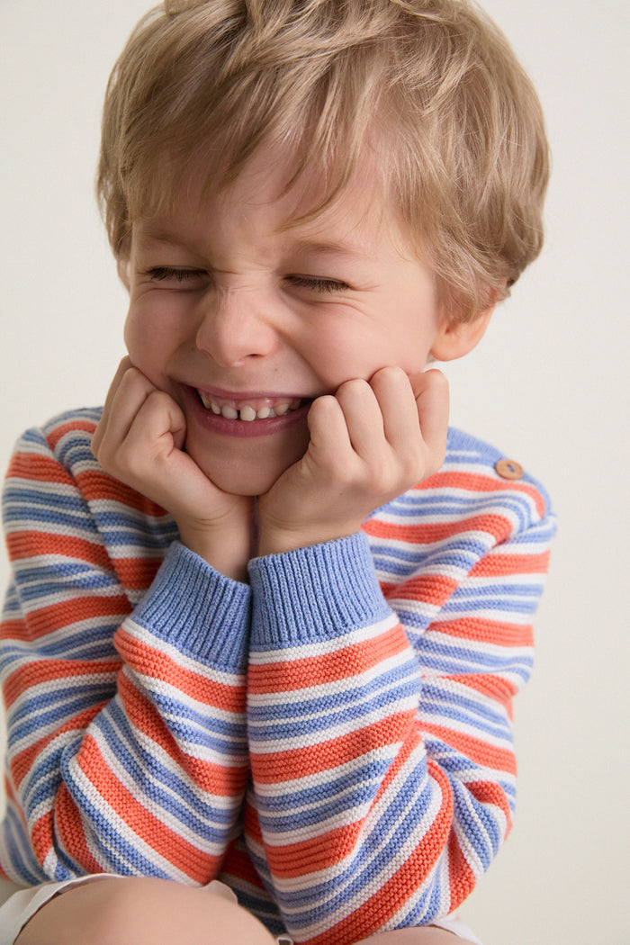 Young child wearing the Coral Stripe Bromo Cotton Jumper, a classic, high-end knit with intarsia stripes and wooden shoulder buttons, showcasing effortless style and fun charm.