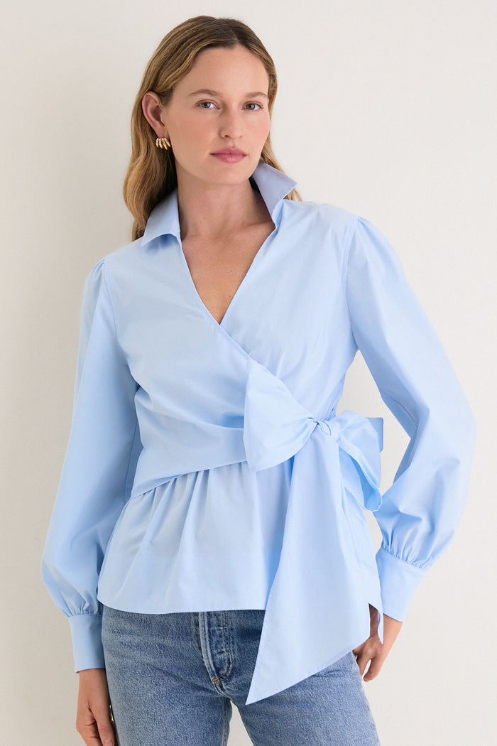 Female model in a classic, effortless French Blue Tie Front Bow Bea Blouse, showcasing its chic wrap design, peplum waist, and elegant self-tie bow.