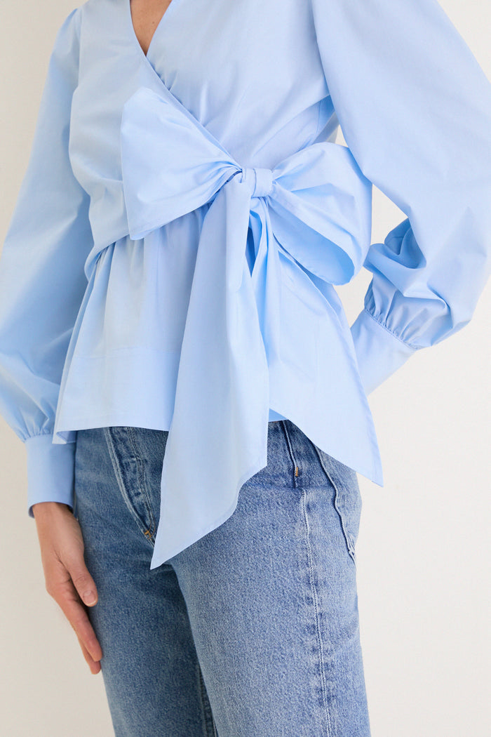 French Blue Tie Front Bow Bea Blouse with classic wrap design, effortless peplum waist, and fun oversized bow, paired stylishly with medium-wash jeans for a high-end look.