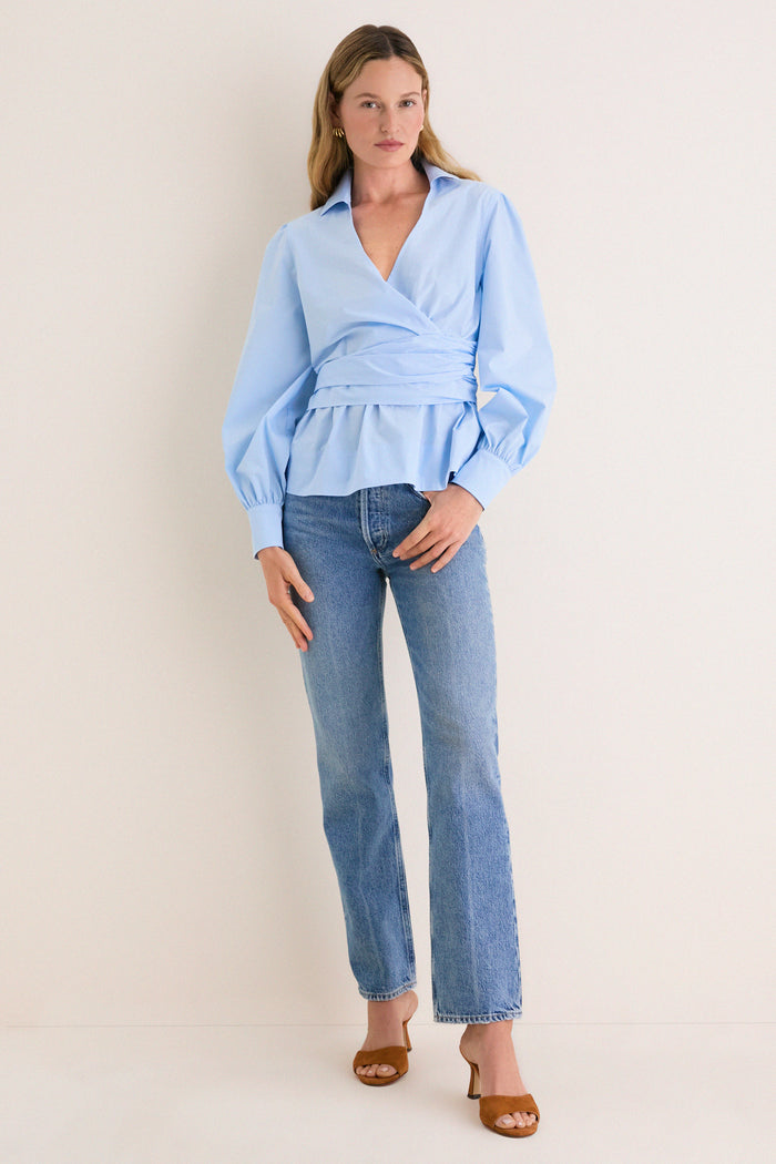 Woman models the classic French Blue Tie Front Bow Bea Blouse, featuring an effortless wrap style and peplum waist, paired with light jeans and tan mule heels.