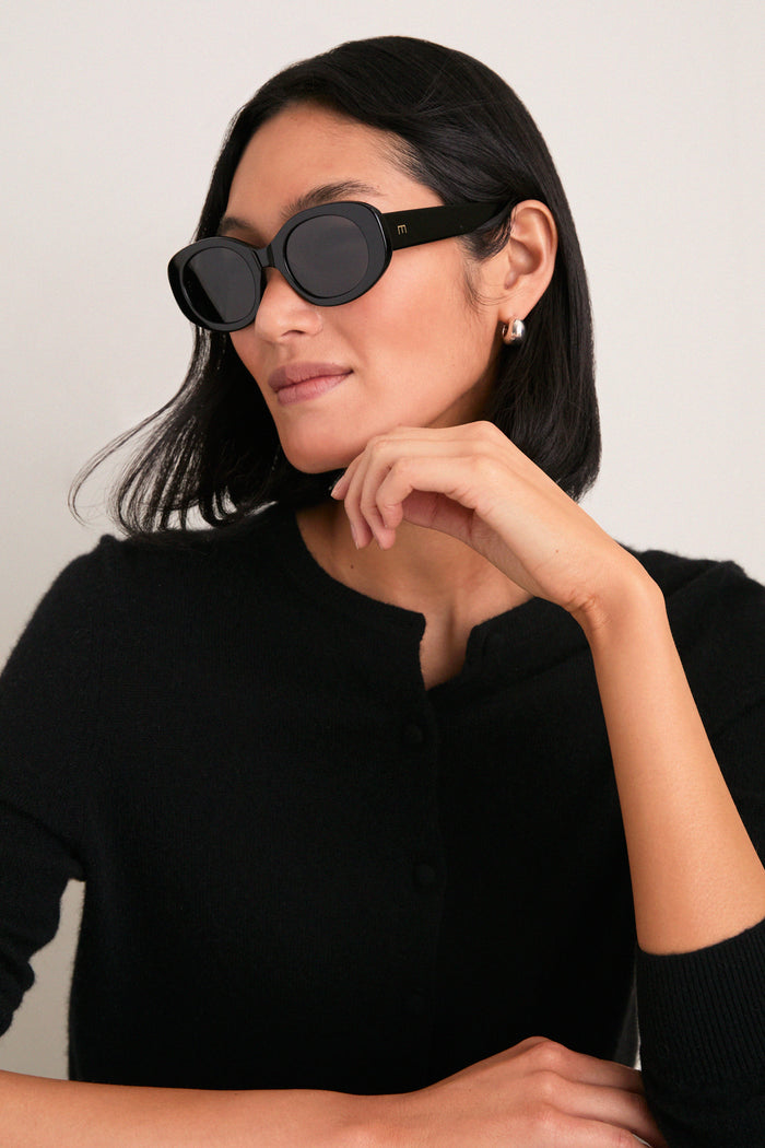Person wearing classic, effortlessly cool Gloss Black Lyna Sunglasses, styled with fitted black cardigan, embodying high-end fashion with fun, bold design by Elisa Johnson.