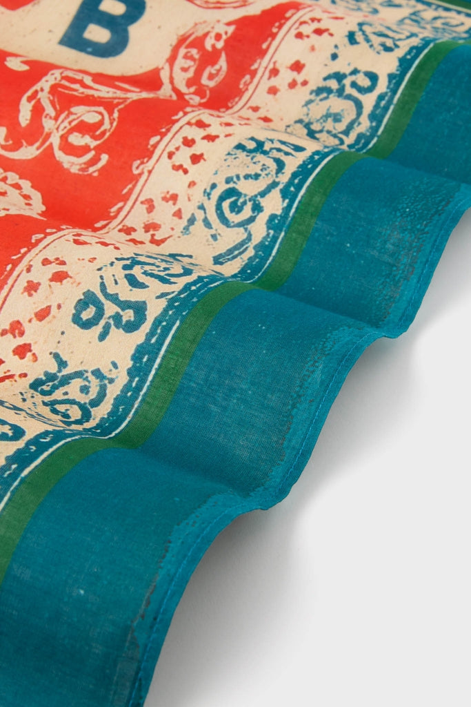 HB x Tuckernuck Texas Strong Scarf features a classic, effortless design with ornate red and blue scroll-work on a natural backdrop, accented by a teal hemmed edge.