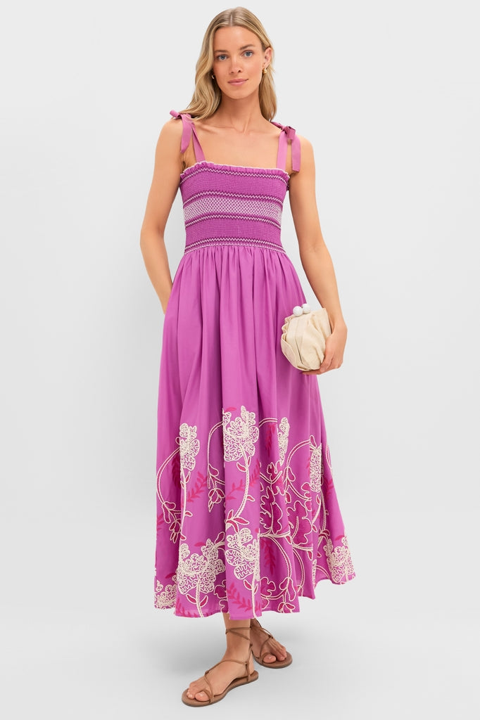 Woman models the Hibiscus Embroidered Scarlett Sundress, featuring a classic smocked bodice, effortless tie-shoulder straps, and fun floral embroidery, paired with a high-end clutch and sandals.