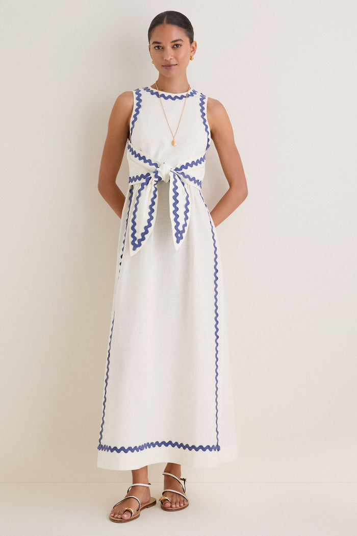 Woman models the classic White Ric Rac Tie-Front Brocks Dress, featuring effortless blue piping detail, complemented by fun gold jewelry and high-end white sandals.
