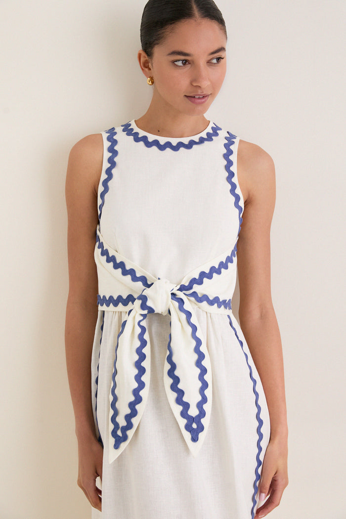 Young woman wearing the classic White Ric Rac Tie-Front Brocks Dress, featuring effortless periwinkle-blue trim and a fun, high-end bow detail at the waist.