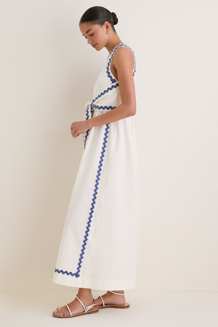 Woman in profile wearing the classic White Ric Rac Tie-Front Brocks Dress, an ankle-length, sleeveless design, exuding effortless elegance with wavy blue ribbon accents.