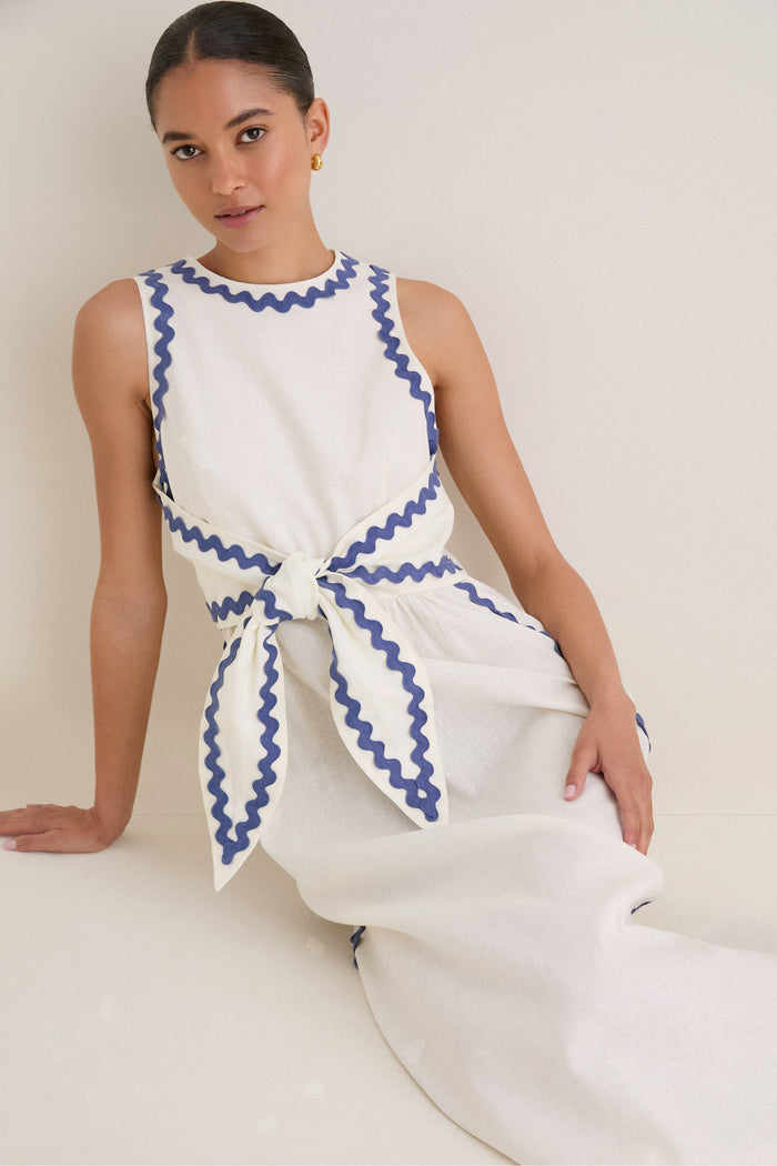 Young woman wearing the classic White Ric Rac Tie-Front Brocks Dress, featuring effortless blue trim details and a fun, high-end sleeveless linen design with a self-tie belt.