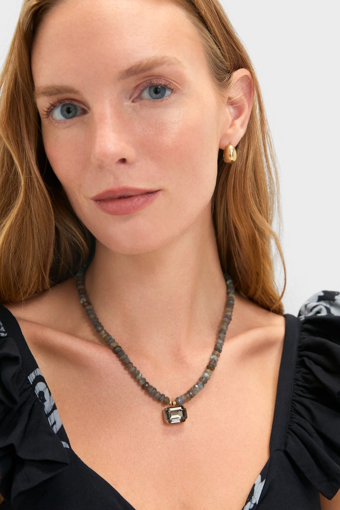 Young woman wearing the classic Labradorite Sidonie Necklace, featuring a faceted gemstone pendant. Her effortless style is complemented by a gold hoop earring and fun, high-end fashion.
