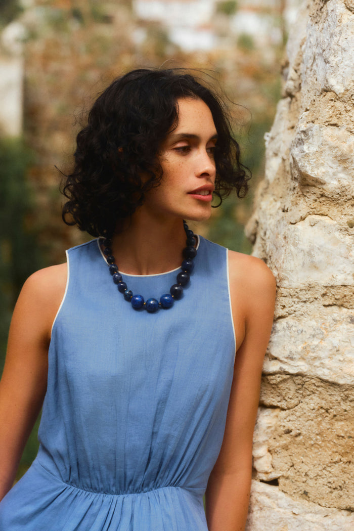 Woman leaning on a stone wall wearing a sleeveless blue dress.