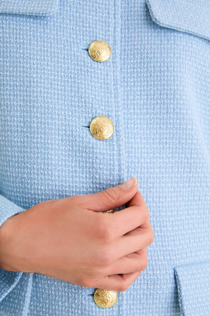 Close-up of Light Blue Tweed Jennings Jacket with gold crest buttons and textured fabric, exemplifying classic style and effortless elegance. Hands are clasped below the shiny buttons.
