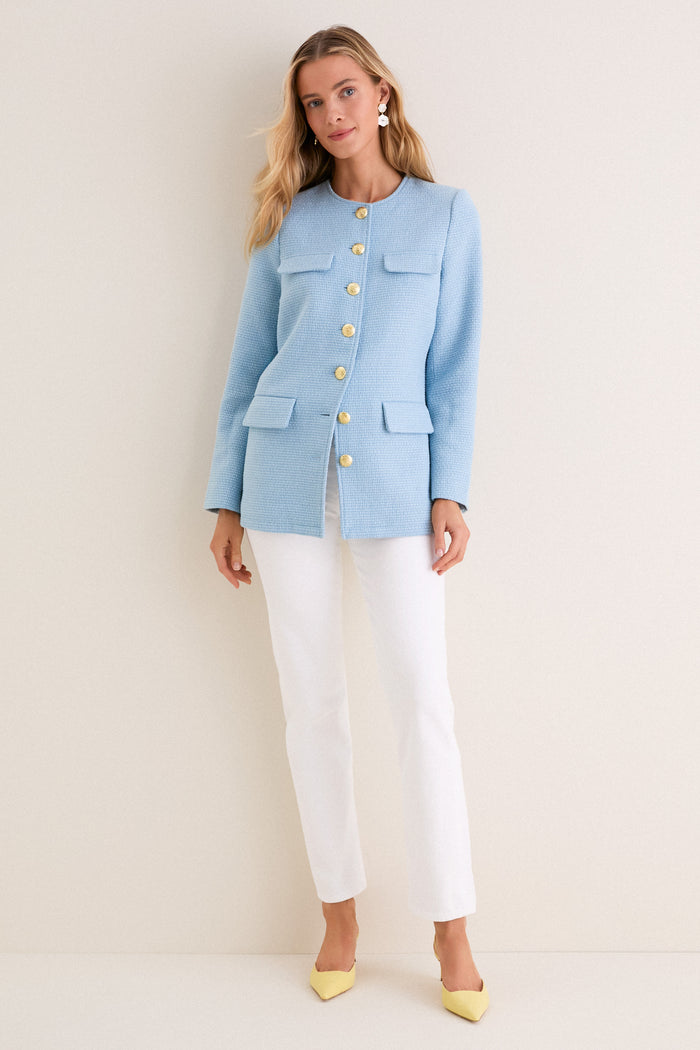 Woman models the Light Blue Tweed Jennings Jacket with gold buttons and flap pockets, styled with white pants and yellow heels for a classic, effortless look.
