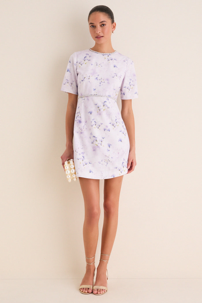 Woman in Lilac Floral Crystal Embellished Floria Mini Dress, clutch and sandals, exuding classic, effortless elegance and fun for elevated daytime events.