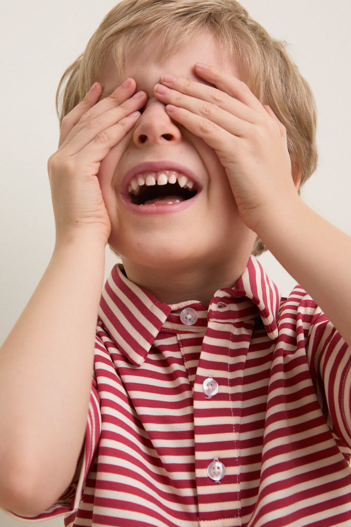 Young boy joyfully plays peek-a-boo in a classic Nantucket Red and Creme Le Club Polo, embodying effortless, fun style with high-end comfort and discrete embroidery.