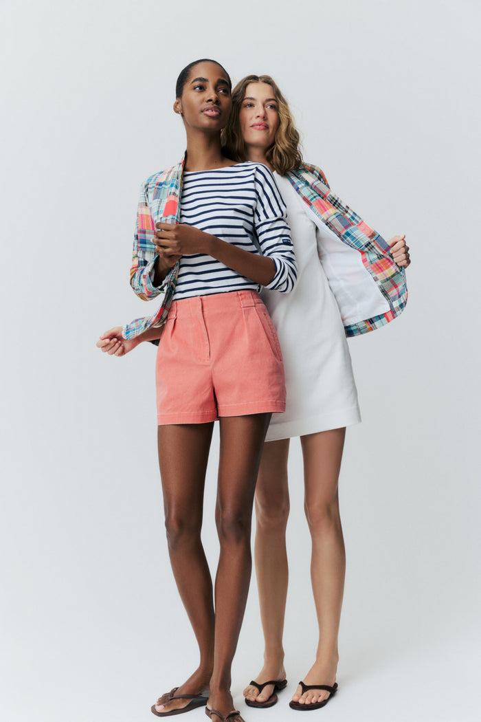 Two women standing together in front of a white backdrop. Woman in the front is wearing a blue and white striped tee and red shorts. Woman behind her is in a white dress and a madras blazer.