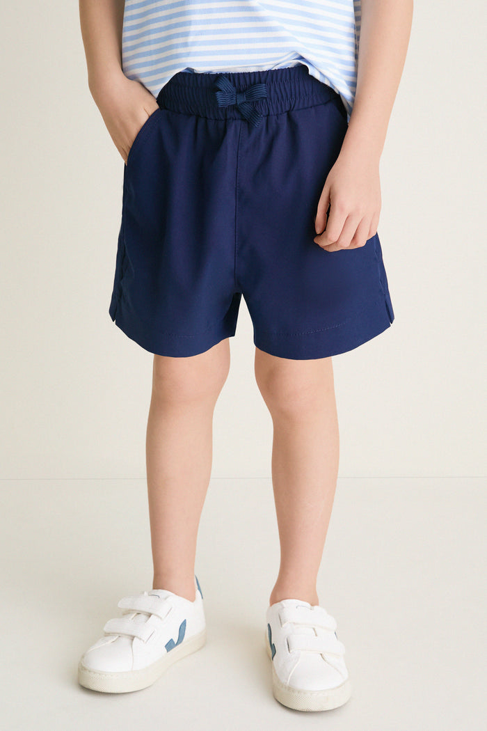 Child's legs in classic Nautical Navy Le Club Shorts, showcasing effortless, high-end style with a tied bow, paired with fun white sneakers.