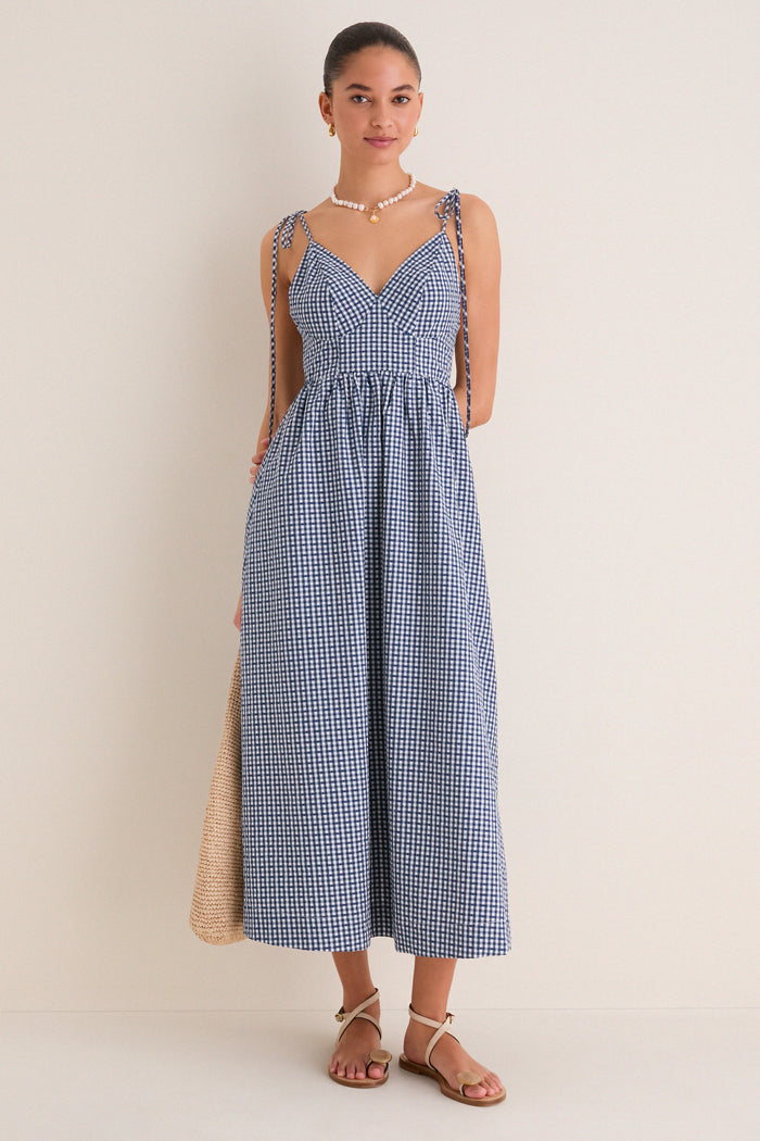 Young woman in a classic, effortless Navy Gingham Sloan Maxi Dress with tie straps, accessorized with pearls and sandals, holding a straw tote, embodying high-end, fun style.