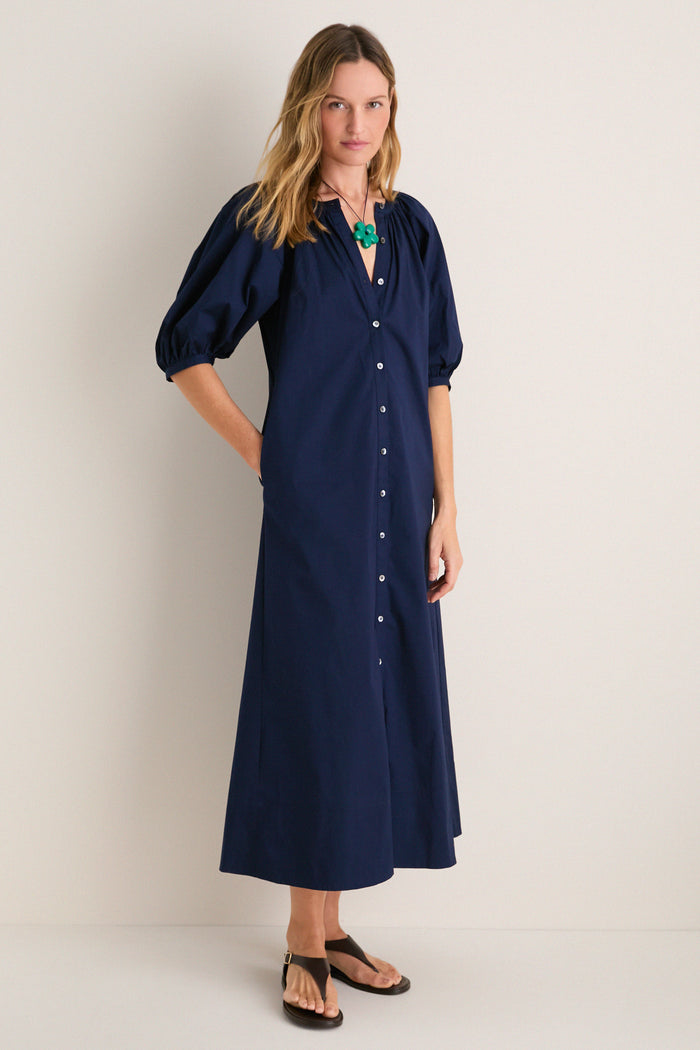 Young woman in a classic, navy Vincent Dress with button-front and puffed sleeves, exuding effortless style, complemented by a bold green pendant and dark brown sandals.