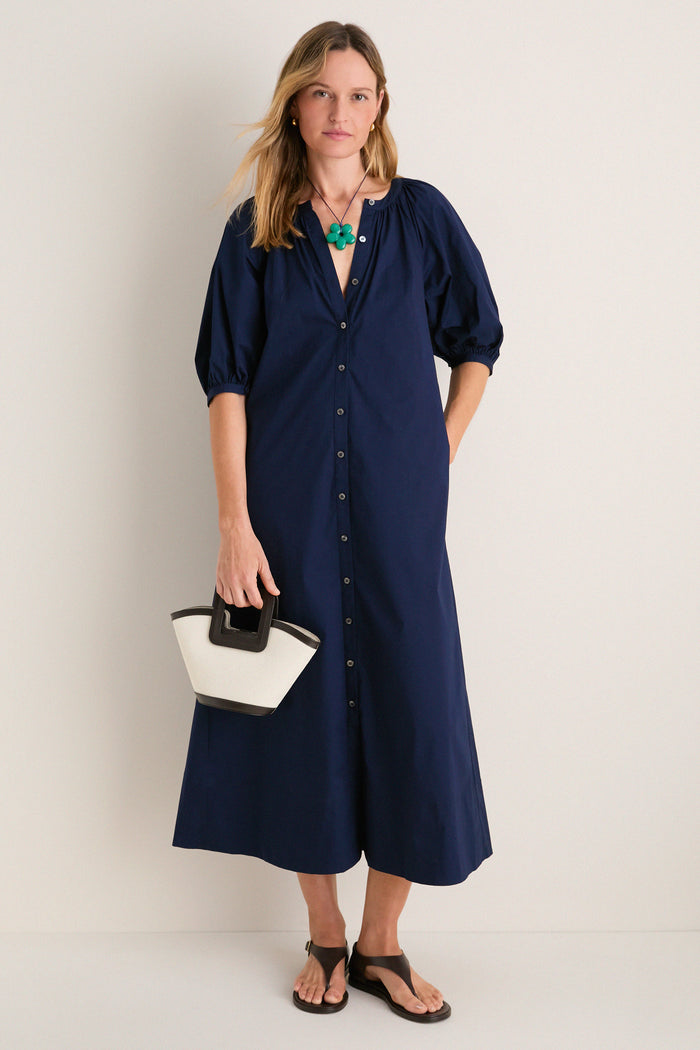 Woman models the classic Navy Vincent Dress, an effortless high-end piece with button-front, balloon sleeves, styled with a fun green pendant and chic accessories.