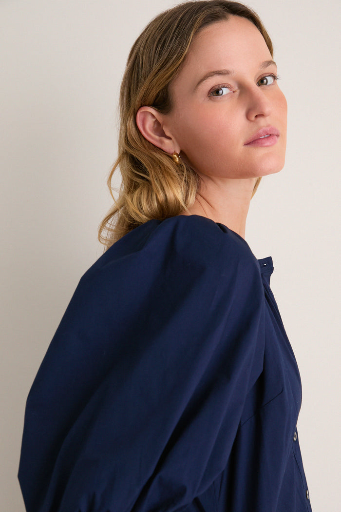 Woman wearing the classic Navy Vincent Dress, featuring a button-down front and balloon sleeves, exuding an effortless, high-end style against a simple backdrop.