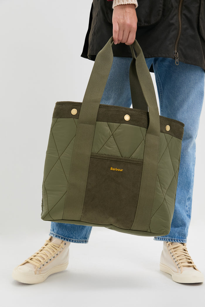 Person holding the classic Olive Healy Tote Bag, featuring effortless quilted design, corduroy pocket with Barbour logo, and high-end webbed handles. Perfect for fun, everyday use.