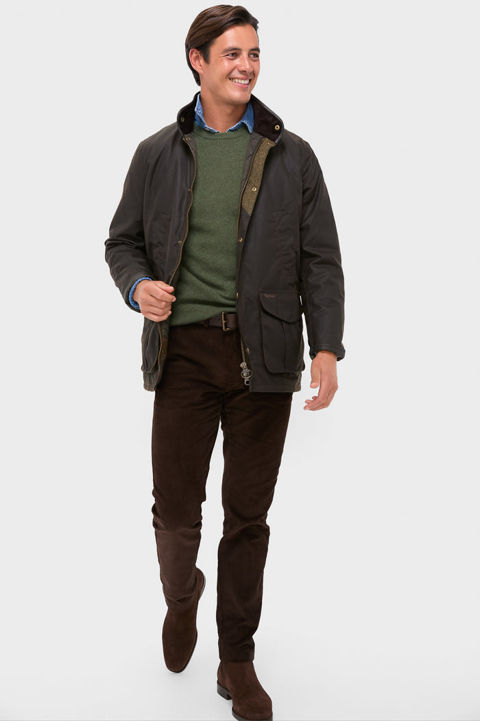 Model wearing the classic Olive Hereford Wax Jacket with a corduroy collar, leather trim, and practical pockets, styled effortlessly with a green sweater and brown corduroy trousers.