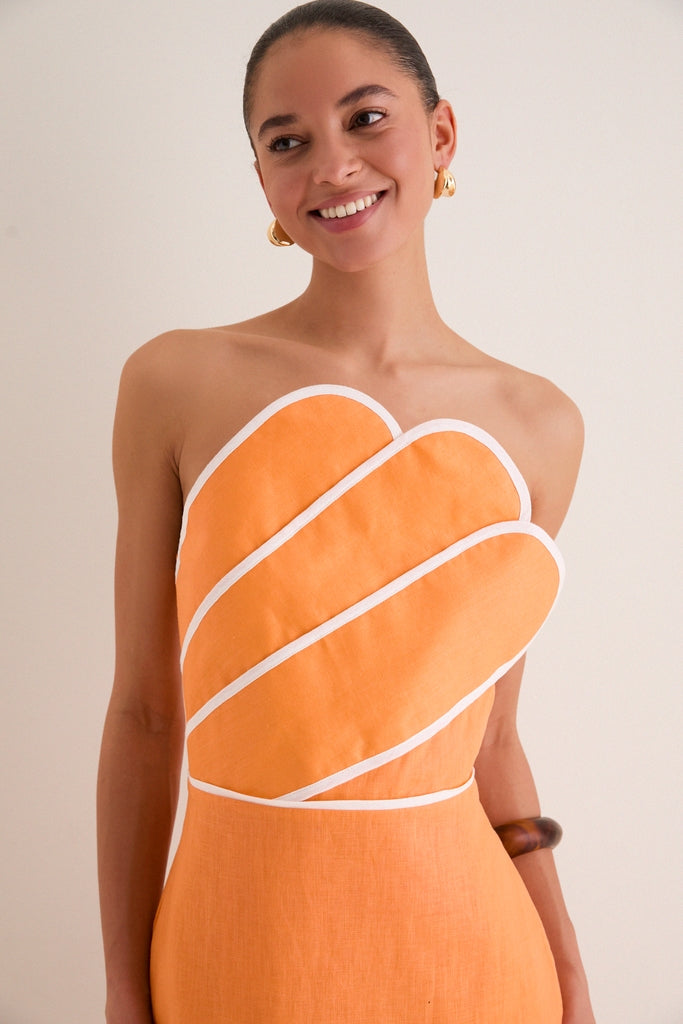 Young woman in an Orange Linen Clementine Maxi Dress with a sculptural, asymmetrical neckline, effortlessly styled with gold hoops and a wooden bangle, exuding classic high-end fun.