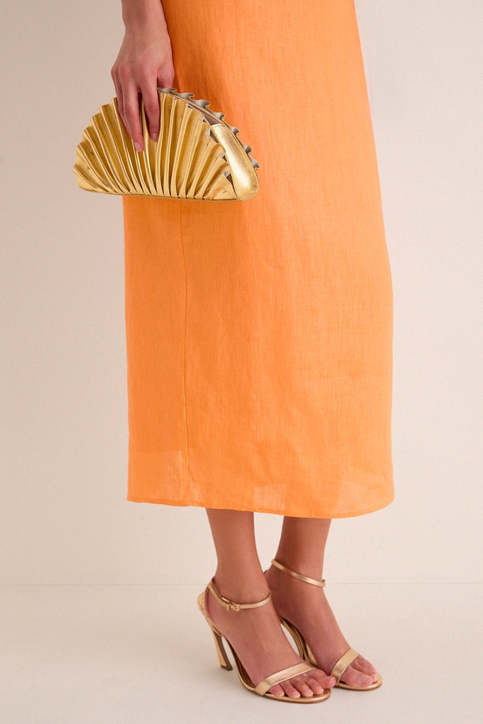 Orange Linen Clementine Maxi Dress with asymmetrical neckline, paired with metallic gold sandals and fan-shaped clutch, embodies classic, effortless style for a fun, high-end look.