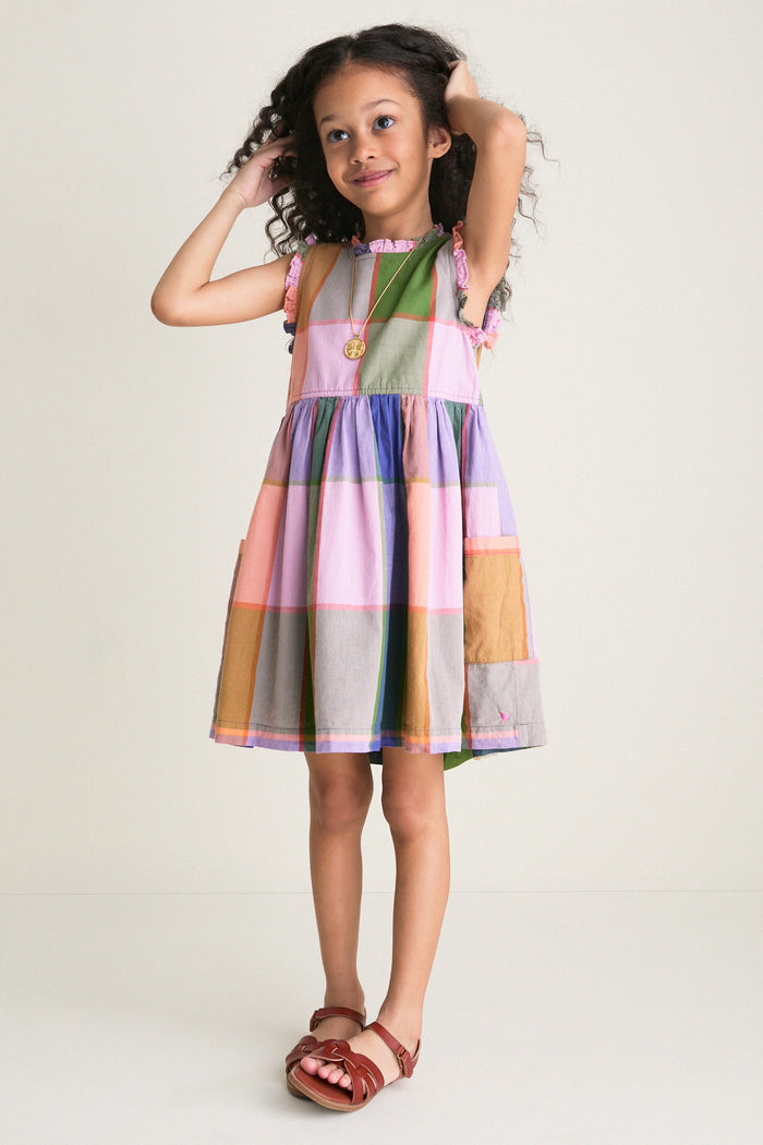 Young girl in a classic, playful Plaid Madras Alyssa Dress, featuring ruffles and patch pockets, embodying effortless style and fun, paired with brown leather sandals.
