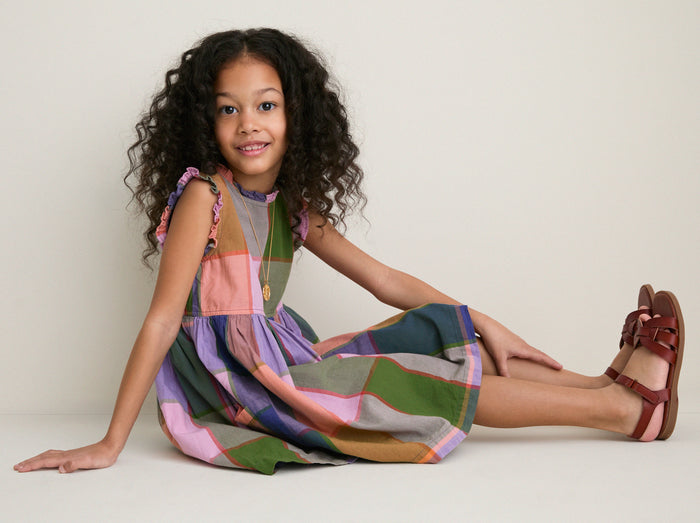 Young girl wears the classic Plaid Madras Alyssa Dress, featuring playful patchwork, ruffle details, and silver buttons, paired with effortless brown sandals and a simple necklace.