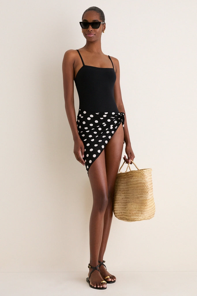 Woman wearing a classic, high-end Polka Dot Noir Bandana as a chic wrap skirt, paired with a black swimsuit, flat sandals, and a woven tote, exuding effortless style.