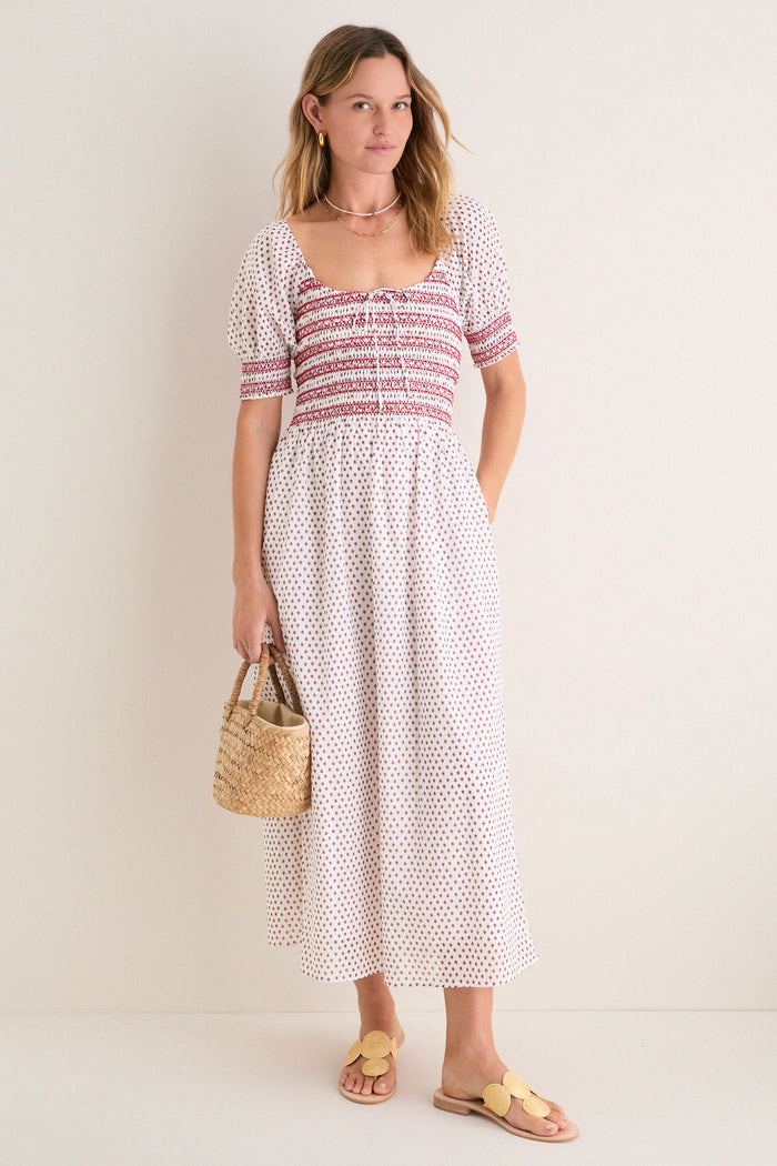 Young woman in a classic, white Madeline Nap Dress with red floral pattern, showcasing effortless elegance. Paired with a woven straw tote and fun raffia slide sandals.