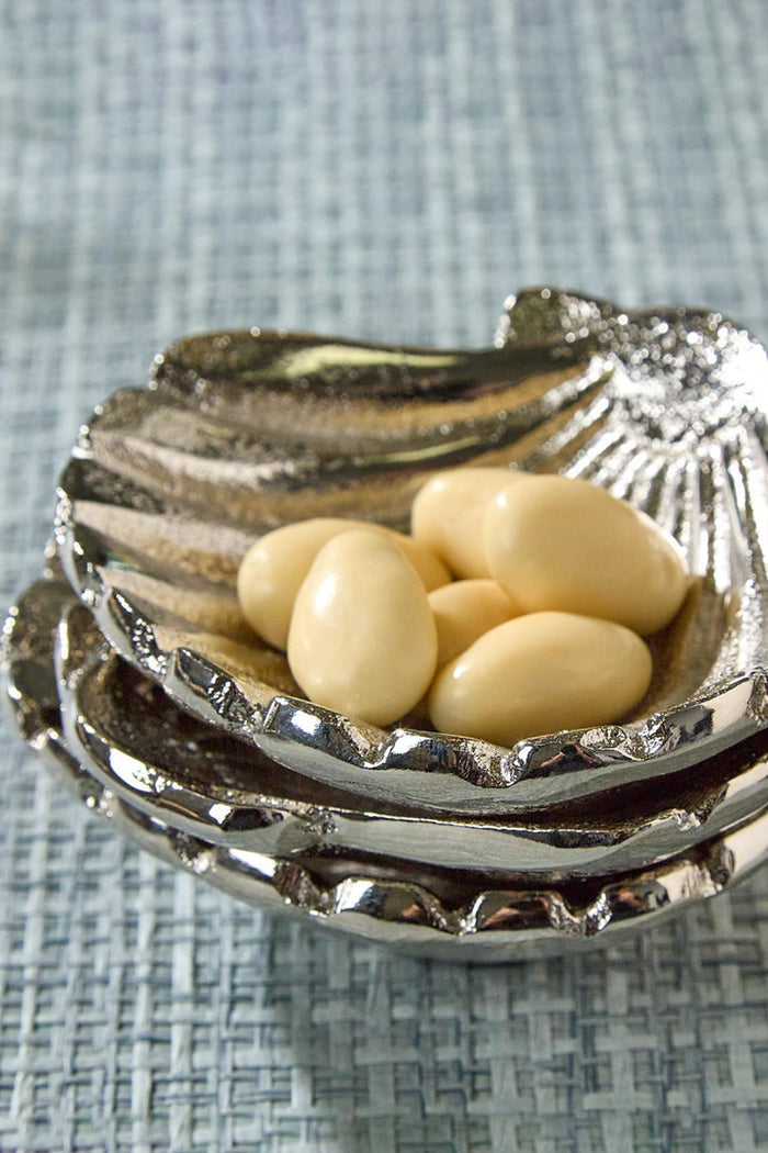 Silver Clam Shell Tidbit Dish holds ivory candy-coated nuts, exuding classic coastal elegance. Crafted from polished aluminum, this high-end piece offers an effortless, fun touch to entertaining.