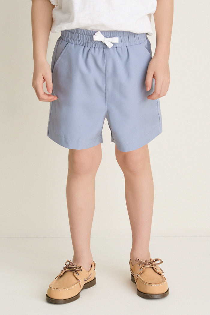 Young child wearing Spanish Slate Le Club Short, showcasing a classic, effortless style with a small white bow and tan boat shoes for a fun, high-end look.