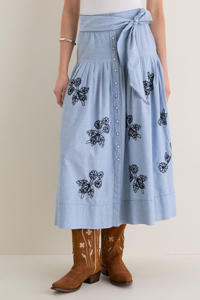 The Chambray Embroidered Highland Skirt features a classic, effortless style with floral embroidery, a flattering dropped waist, and a fun self-tie, paired with high-end cowboy boots.