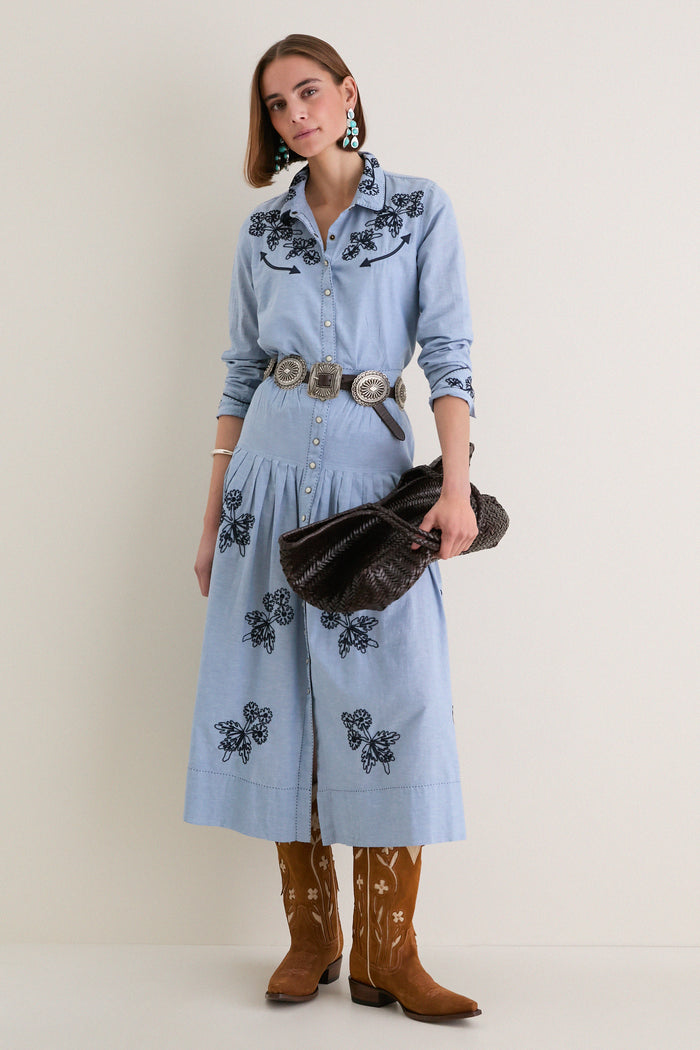 Woman wearing The Chambray Embroidered Highland Skirt with navy floral embroidery, paired with a silver concho belt, turquoise earrings, and cowboy boots for a classic, effortless look.