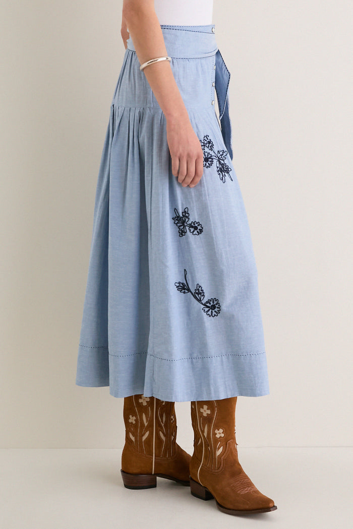 The Chambray Embroidered Highland Skirt, featuring classic navy floral embroidery and pearl snaps, styled with cowboy boots for an effortless, high-end, and fun look.